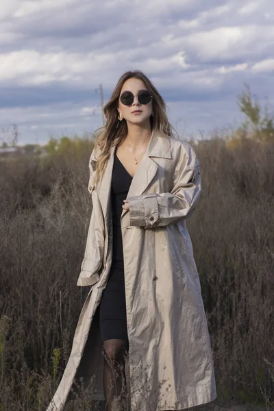brunette in fashionable trench coat, black dress and sunglasses in the field. autumn, yellow leaves, blue sky