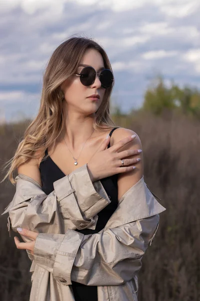 Brunette in modieuze trench jas, zwarte jurk en zonnebril in het veld. herfst, gele bladeren, blauwe lucht — Stockfoto