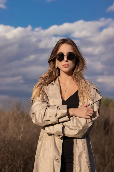 Brunette in modieuze trench jas, zwarte jurk en zonnebril in het veld. herfst, gele bladeren, blauwe lucht — Stockfoto