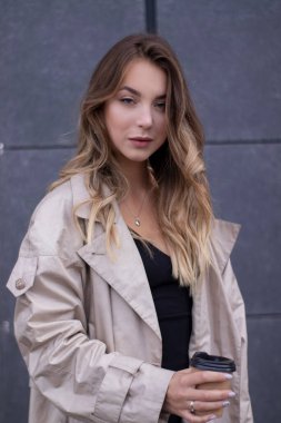 şık trençkotlu ve siyah elbiseli, kağıt bardaklı, ince, genç bir esmer.