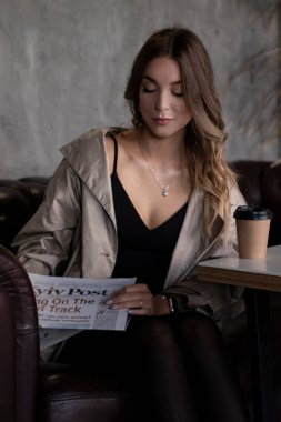 Gri çatı katındaki trençkotlu esmer, kağıt bardak ve gazeteyle deri koltukta. şehir yaşam tarzı