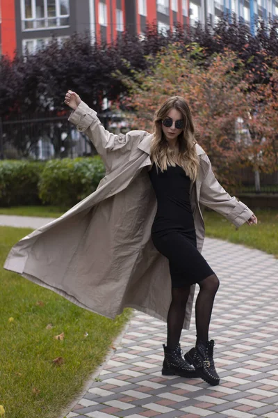 Atraente slim jovem morena na trincheira da moda, vestido preto e óculos de sol no parque. Outono, folhas amarelas — Fotografia de Stock