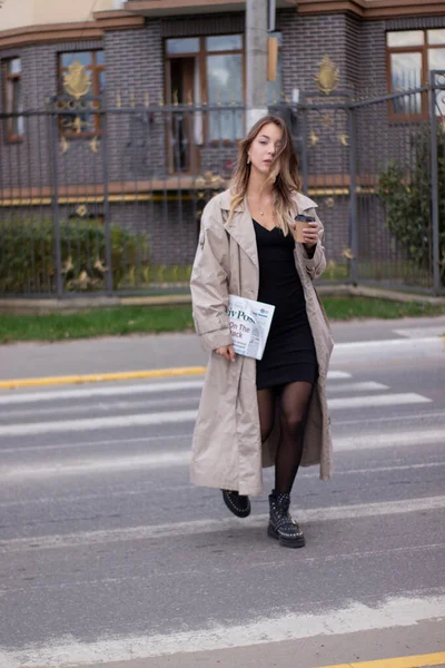 Belle jeune brune mince en trench-coat à la mode et robe noire traversent un passage piéton — Photo