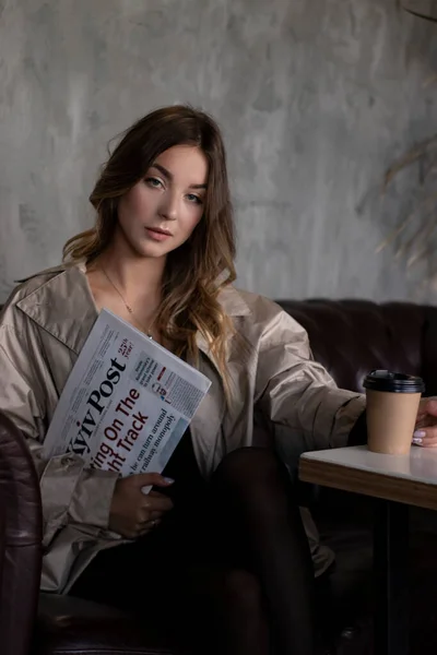 Atrakcyjna szczupła brunetka w płaszczu w szarym pokoju na poddaszu na skórzanej kanapie z papierową filiżanką i gazetą. miejski styl życia — Zdjęcie stockowe