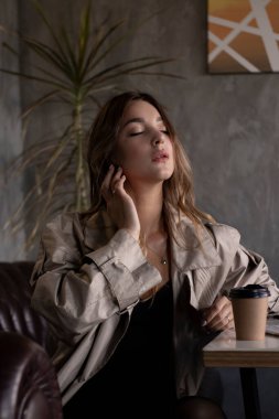 Gri çatı katındaki trençkotlu esmer, deri koltukta, kağıt bardakla. şehir yaşam tarzı kavramı