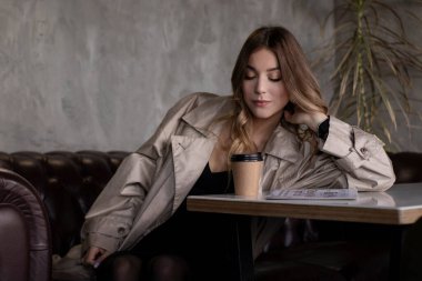 Gri çatı katındaki trençkotlu esmer, kağıt bardak ve gazeteyle deri koltukta. şehir yaşam tarzı