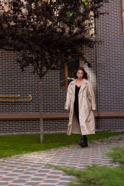 Moda trençkotlu ve siyah elbiseli çekici, zayıf esmer. Sonbahar, sarı yapraklar. tuğla bina