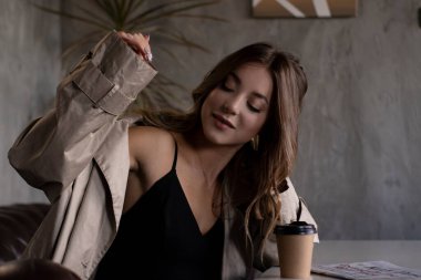 Gri çatı katındaki trençkotlu esmer, deri koltukta, kağıt bardakla. şehir yaşam tarzı kavramı