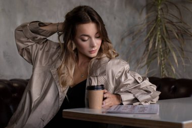 Gri çatı katındaki trençkotlu esmer, kağıt bardak ve gazeteyle deri koltukta. şehir yaşam tarzı