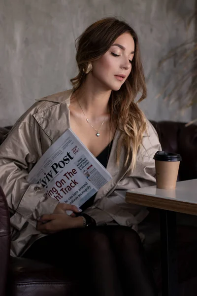 Jolie brune mince en trench coat dans la chambre grise loft sur canapé en cuir avec tasse en papier et journal. mode de vie urbain — Photo