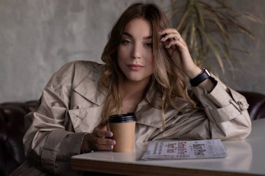 Gri çatı katındaki trençkotlu esmer, kağıt bardak ve gazeteyle deri koltukta. şehir yaşam tarzı