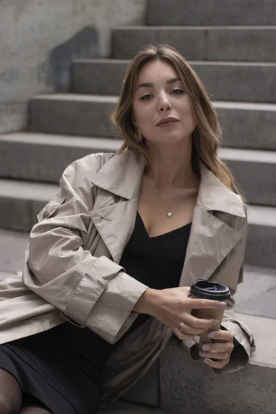 Atractiva morena en trinchera de moda y vestido negro sentado en escalera de hormigón gris con café taza de papel marrón —  Fotos de Stock