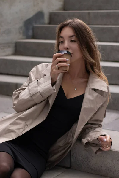 Jolie brune dans une tranchée à la mode et robe noire assise sur un escalier en béton gris avec tasse en papier brun café — Photo