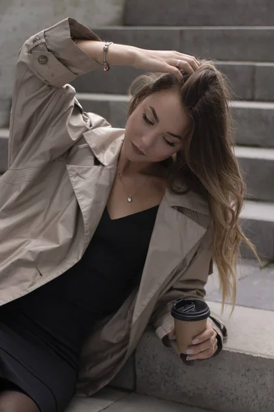 Aantrekkelijke brunette in modieuze trench en zwarte jurk zitten op grijze betonnen trap met bruine papieren kopje koffie — Stockfoto
