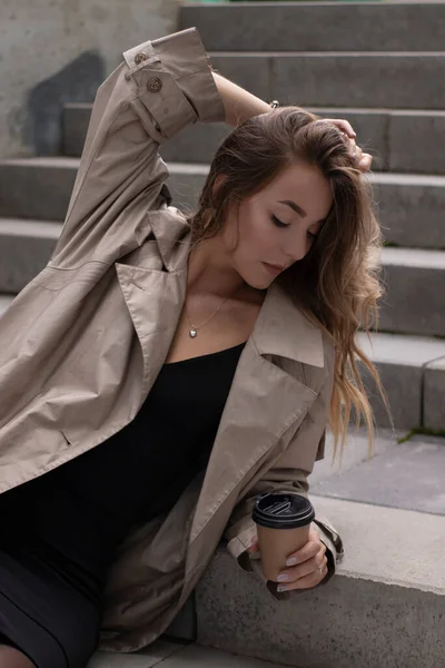 Jolie brune dans une tranchée à la mode et robe noire assise sur un escalier en béton gris avec tasse en papier brun café — Photo