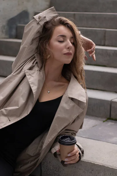 Jolie brune dans une tranchée à la mode et robe noire assise sur un escalier en béton gris avec tasse en papier brun café — Photo