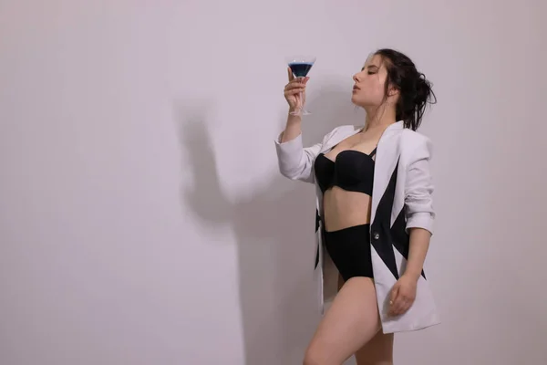 Σέξι ελκυστική μελαχρινή με μαύρα εσώρουχα και λευκό και μαύρο κομψό σακάκι με ένα μπλε κοκτέιλ σε γκρι δωμάτιο. αλκοόλ, κόμμα έννοια — Φωτογραφία Αρχείου