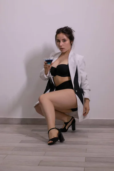 Σέξι ελκυστική μελαχρινή με μαύρα εσώρουχα και λευκό και μαύρο κομψό σακάκι με ένα μπλε κοκτέιλ σε γκρι δωμάτιο. αλκοόλ, κόμμα έννοια — Φωτογραφία Αρχείου