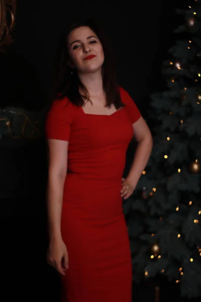 Σέξι ελκυστική μελαχρινή με κόκκινο κοκαλιάρικο φόρεμα κοντά σε χριστουγεννιάτικο δέντρο. σκοτεινή φωτογραφία. Φωτογράφηση στούντιο. συναισθηματική ηθοποιός. — Φωτογραφία Αρχείου