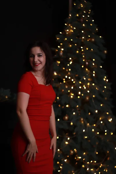 Sexy aantrekkelijke brunette in rood mager jurk in de buurt van een kerstboom. donkere foto. Studioshoot. emotionele actrice. — Stockfoto