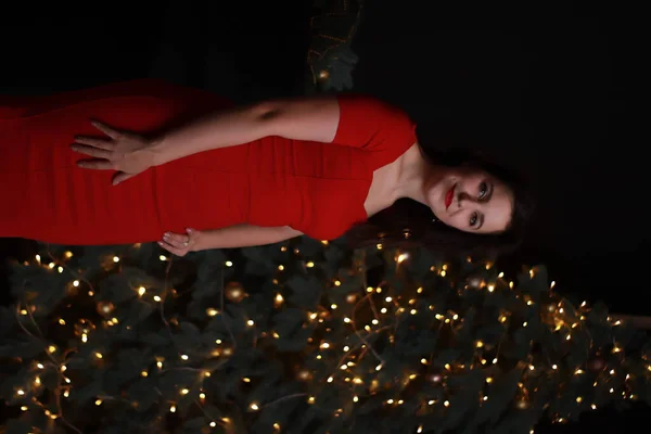 Sexy aantrekkelijke brunette in rood mager jurk in de buurt van een kerstboom. donkere foto. Studioshoot. emotionele actrice. — Stockfoto