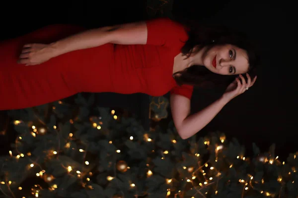 Sexy aantrekkelijke brunette in rood mager jurk in de buurt van een kerstboom. donkere foto. Studioshoot. emotionele actrice. — Stockfoto