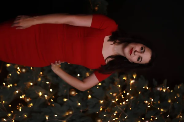 クリスマスツリーの近くの赤いスキニードレスのセクシーな魅力的なブルネット。暗い写真だ。スタジオ撮影。感情的な女優. — ストック写真