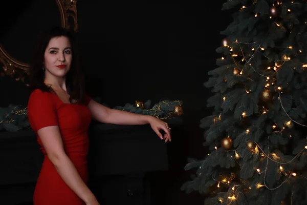 Sexy morena atractiva en vestido rojo flaco cerca de un árbol de Navidad. foto oscura. sesión de estudio. actriz emocional. —  Fotos de Stock