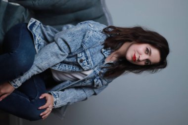 Kotlu ve kot ceketli çekici esmer gri koltukta oturuyor. Stüdyo çekimi