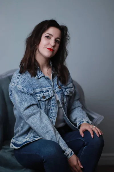 Aantrekkelijke brunette in jeans en spijkerjasje zittend op grijze stoel. studio opname — Stockfoto