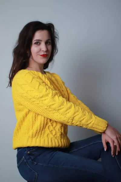 Atractiva morena en jeans y suéter de punto amarillo sobre fondo hrey. sesión de estudio —  Fotos de Stock