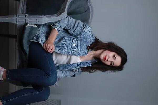 穿着牛仔裤和牛仔夹克的漂亮的黑发女人坐在灰色的椅子上。工作室拍摄 — 图库照片