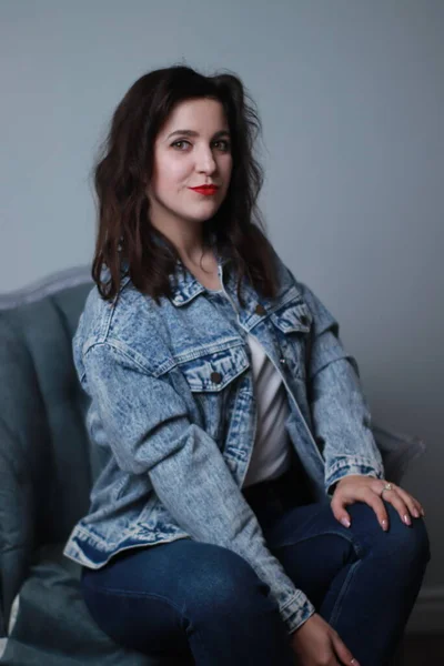 Attractive brunette in jeans and denim jacket sitting on grey chair. studio shoot — Stock Photo, Image