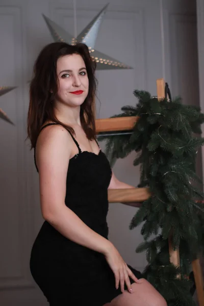 Sexy morena atractiva en vestido negro flaco en escalera envuelta con árbol de Navidad decorativo. sesión de estudio. año nuevo tema —  Fotos de Stock