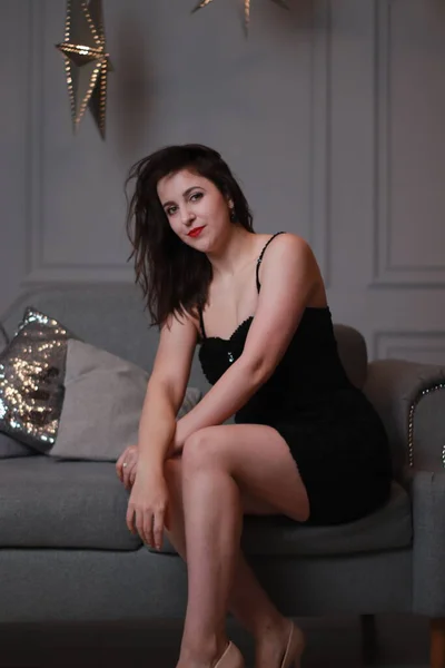 Sexy morena atractiva en vestido negro flaco en un sofá gris en una habitación gris con grandes estrellas decorativas. sesión de estudio —  Fotos de Stock