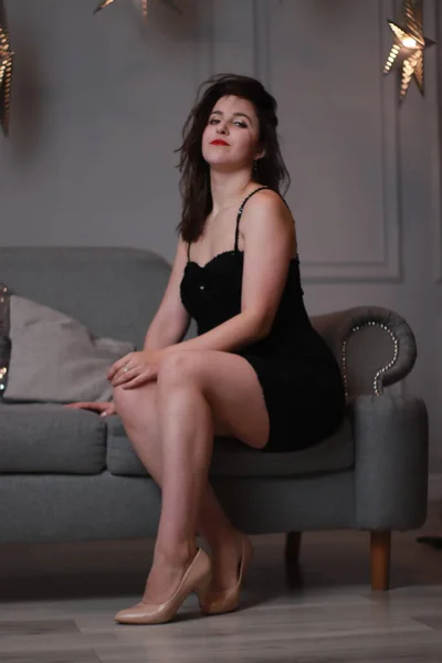 Sexy morena atractiva en vestido negro flaco en un sofá gris en una habitación gris con grandes estrellas decorativas. sesión de estudio —  Fotos de Stock