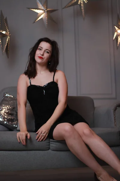 Σέξι ελκυστική μελαχρινή σε μαύρο κοκαλιάρικο φόρεμα σε γκρι καναπέ σε γκρι δωμάτιο με διακοσμητικά μεγάλα αστέρια. φωτογράφιση στούντιο — Φωτογραφία Αρχείου