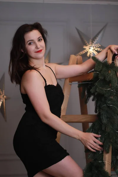 Sexy morena atractiva en vestido negro flaco en escalera envuelta con árbol de Navidad decorativo. sesión de estudio. año nuevo tema —  Fotos de Stock