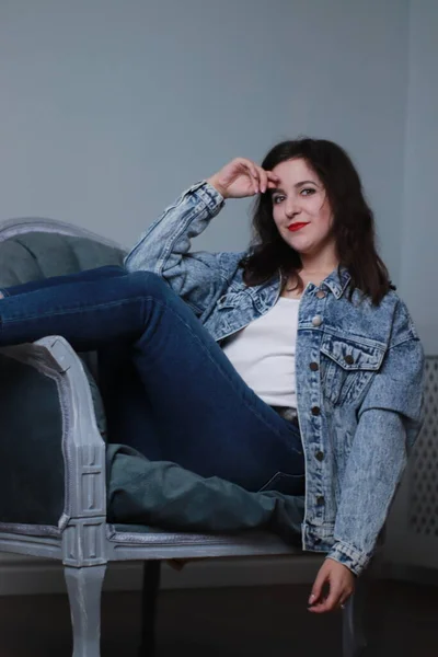 Morena atraente em jeans e casaco de ganga sentado na cadeira cinza. estúdio atirar — Fotografia de Stock