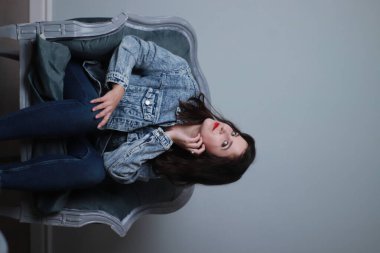 Kotlu ve kot ceketli çekici esmer gri koltukta oturuyor. Stüdyo çekimi