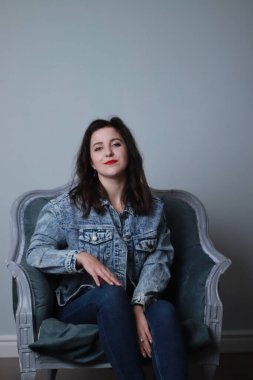 Kotlu ve kot ceketli çekici esmer gri koltukta oturuyor. Stüdyo çekimi