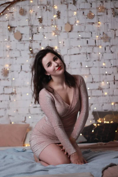 Σέξι ελκυστική μελαχρινή σε μπεζ φόρεμα κάθεται σε ένα κρεβάτι. Φωτογράφηση στούντιο. κόμμα, έννοια εορτασμού — Φωτογραφία Αρχείου