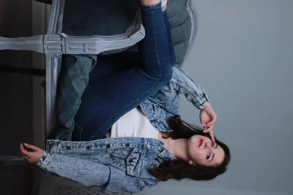 Morena atraente em jeans e casaco de ganga sentado na cadeira cinza. estúdio atirar — Fotografia de Stock