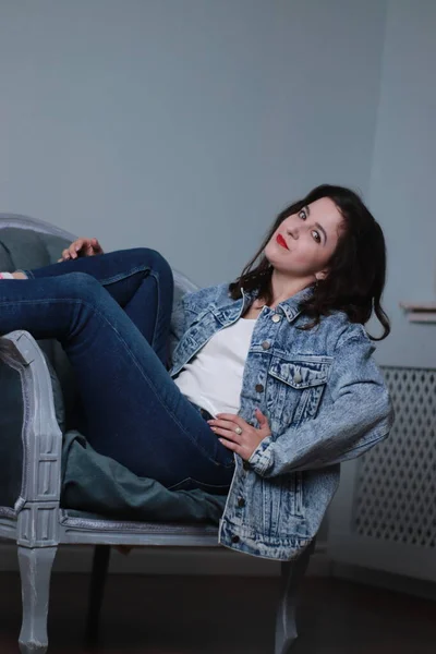 Atractiva morena en jeans y chaqueta de mezclilla sentada en silla gris. sesión de estudio — Foto de Stock