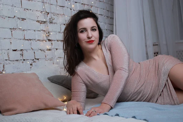 Sexy attraktive Brünette in beigem Kleid auf einem Bett liegend. Studioaufnahmen. Party, Festkonzept — Stockfoto
