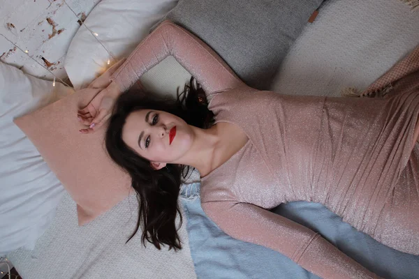 ベージュドレスのセクシーな魅力的なブルネットがベッドの上に横たわっています。スタジオ撮影。パーティーやお祝いのコンセプト — ストック写真