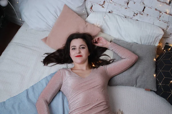 Sexy atraktivní brunetka v béžových šatech ležící na posteli. Studio natáčení. párty, koncepce oslav — Stock fotografie