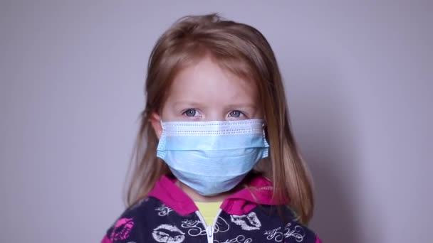 Niña rubia molesta con una máscara médica azul. se quita la máscara. en un suéter rosa. covid 19 pandemia. niños en aislamiento. Imágenes de FullHD. — Vídeo de stock