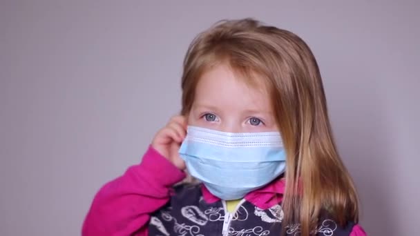 穿着蓝色医疗面具的金发小女孩很不高兴。摘下面具。穿着粉色毛衣。covid 19 pandemia 。被隔离的孩子FullHD视频. — 图库视频影像