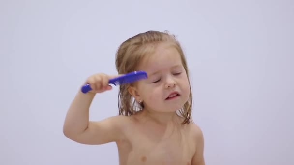 Gadis pirang kecil yang lucu menyisir rambutnya dengan sisir biru pada latar belakang putih. perawatan rambut. Orang tua. Kemerdekaan. Rekaman full-HD — Stok Video